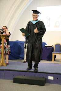 A tall white man in graduation gown giving thumbs up to camera